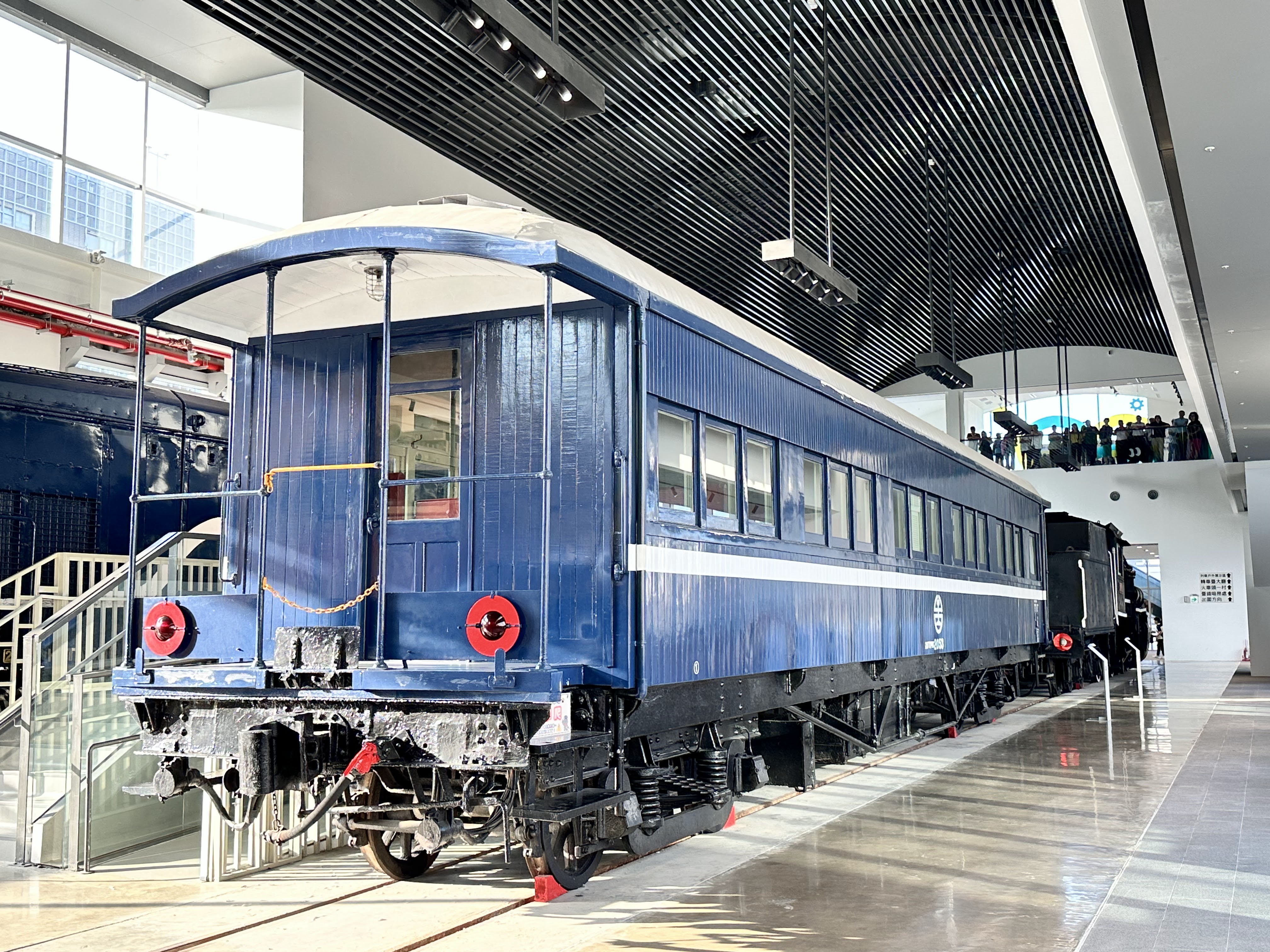 一節復古藍色火車車廂，停放在室內展覽空間內，旁邊有樓梯和參觀民眾。