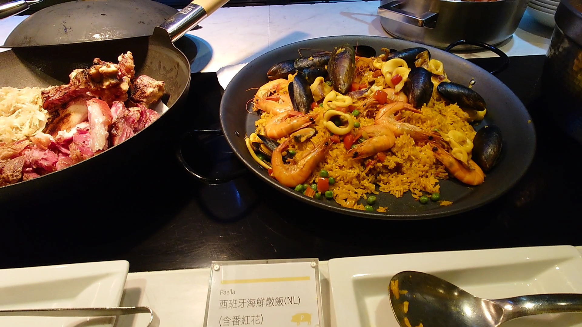 這張圖片展示自助餐檯上的西班牙海鮮燉飯（Paella），飯裡配有鮮蝦、淡菜、花枝和蔬菜，色彩豐富，左邊還有烤豬腳和酸菜。圖片底部可以看到菜名標示，沒有明顯浮水印。