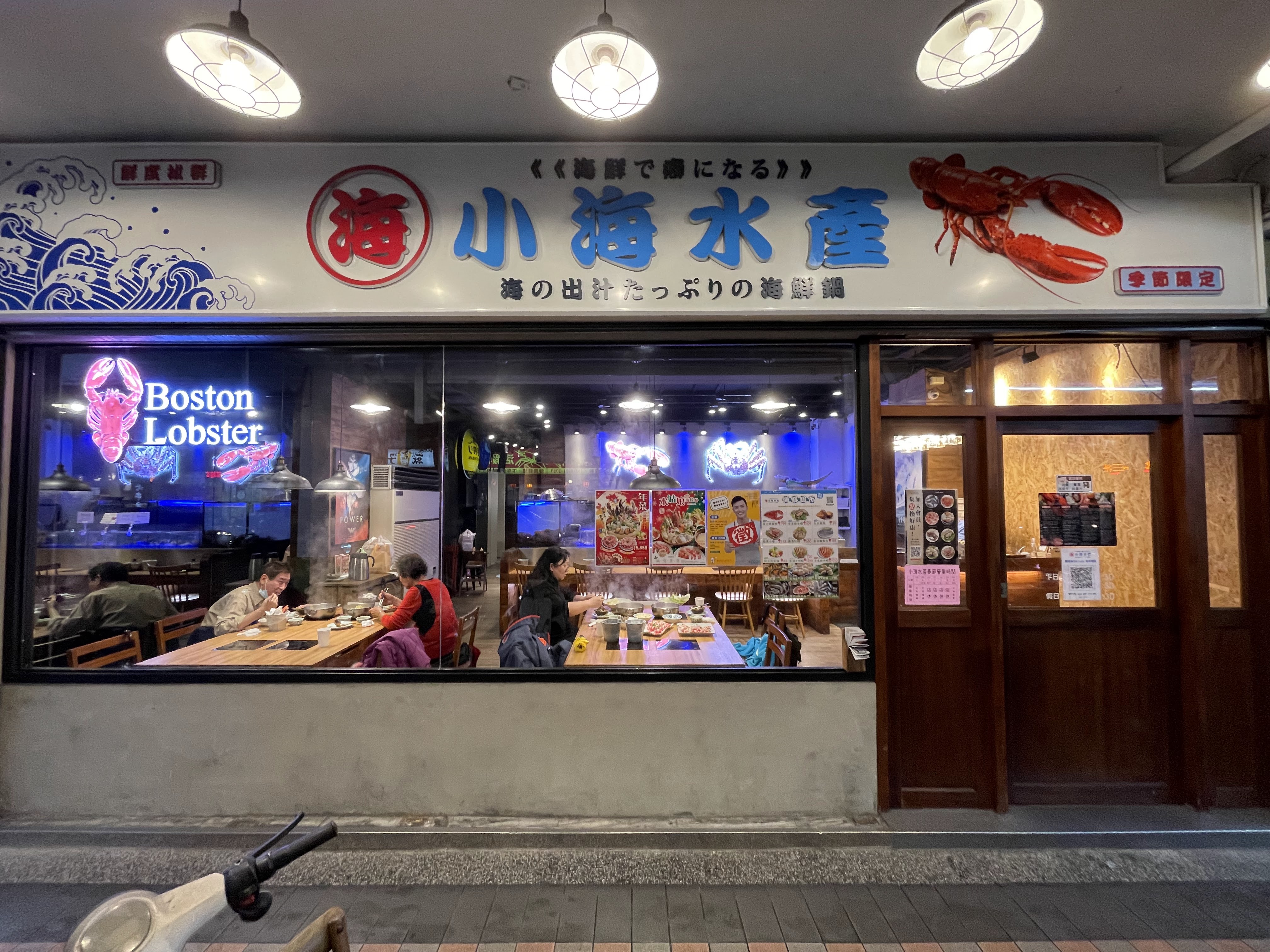 這是一家主打波士頓龍蝦與海鮮火鍋料理的餐廳外觀,招牌上有大龍蝦圖案和中日文名稱,玻璃窗內能看到用餐客人,整體氣氛溫馨明亮,未見明顯浮水印。