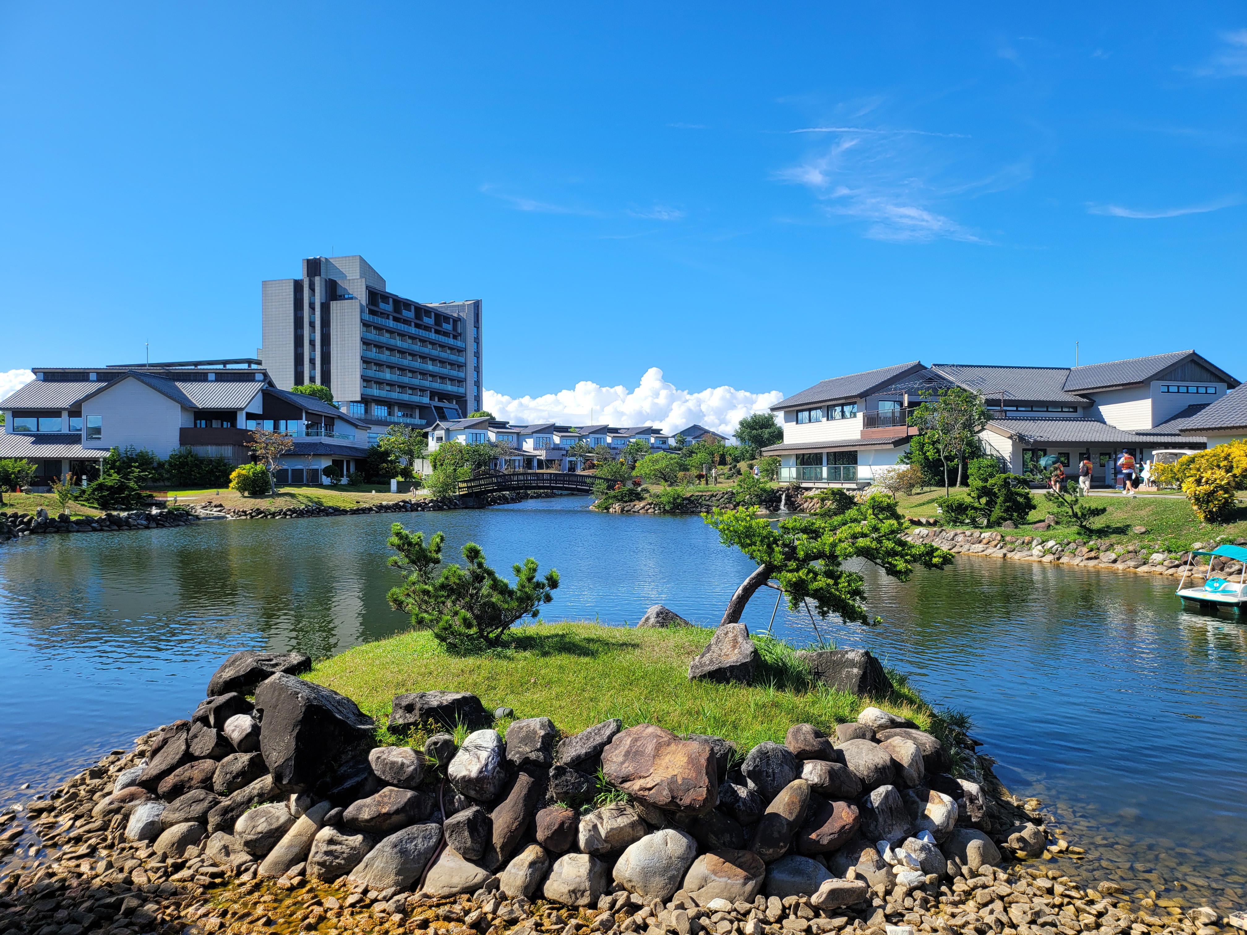 這張圖片展示了一個有池塘和小島的日式庭園,周圍有傳統風格建築和現代高樓,藍天白雲下景色優美。圖片沒有浮水印。