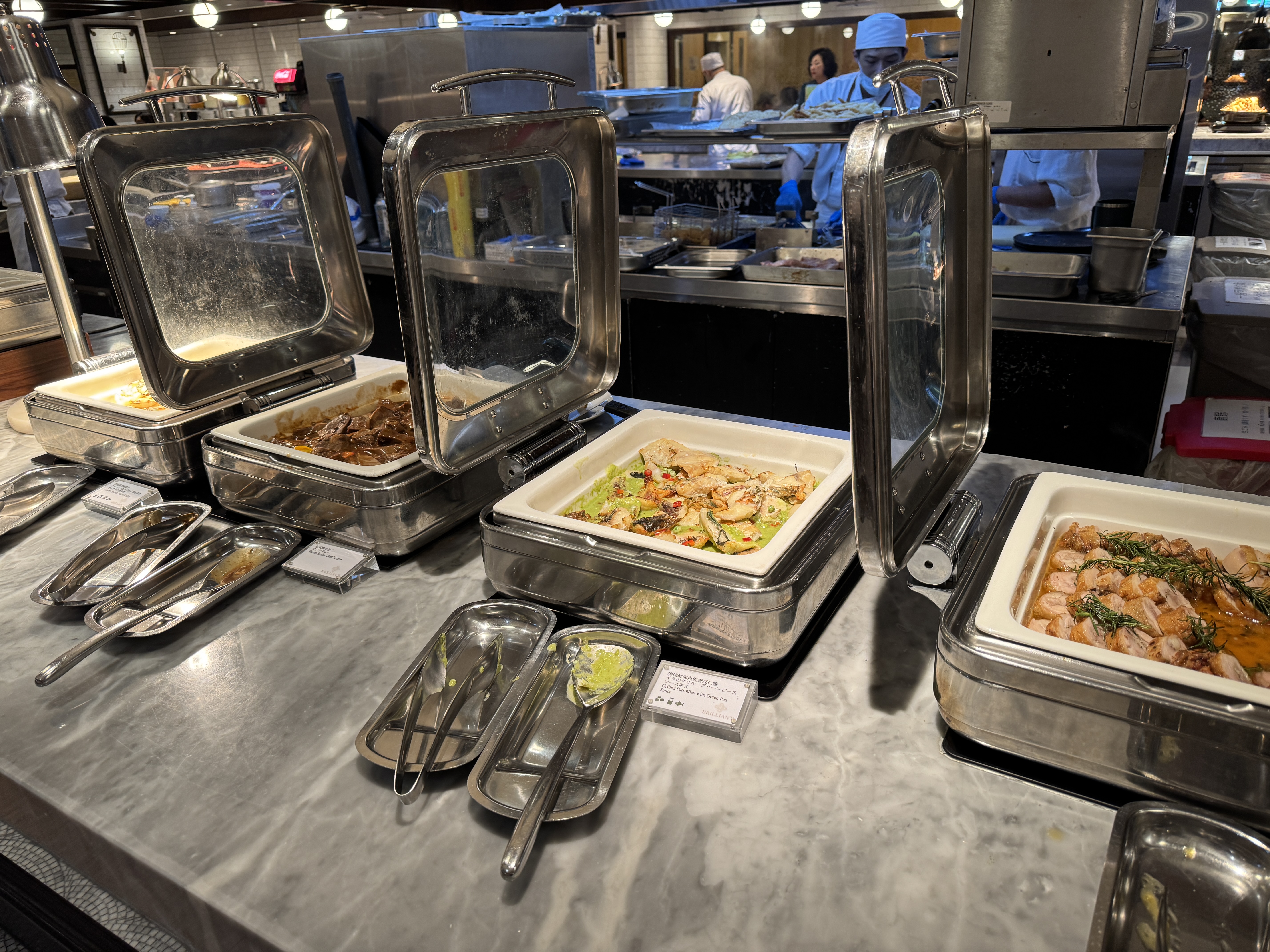 這張圖片主要是自助餐台,擺放著多種熱食菜餚,包括炒蔬菜、肉類等。後方有廚師正在廚房工作,整體環境明亮、衛生。圖片上未見明顯浮水印。