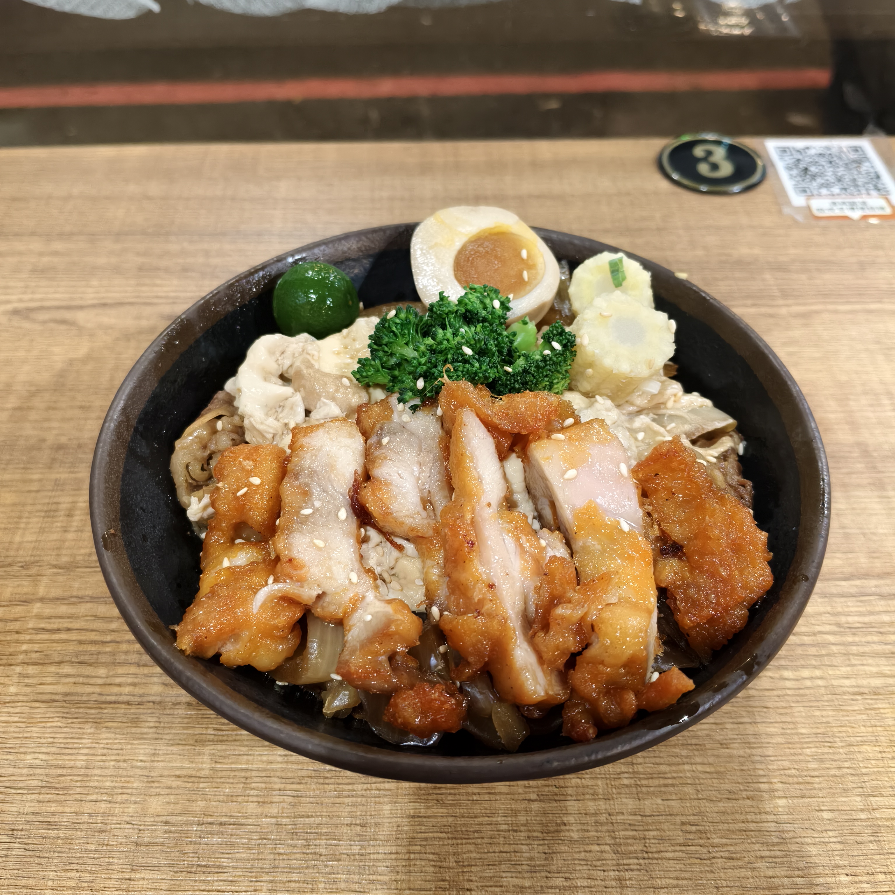 這是一碗日式蓋飯，配有炸雞塊、溏心蛋、青檸、豆皮、花椰菜和天婦羅，全部擺放在白飯上。碗中食材豐富且色彩鮮明，圖片無浮水印。