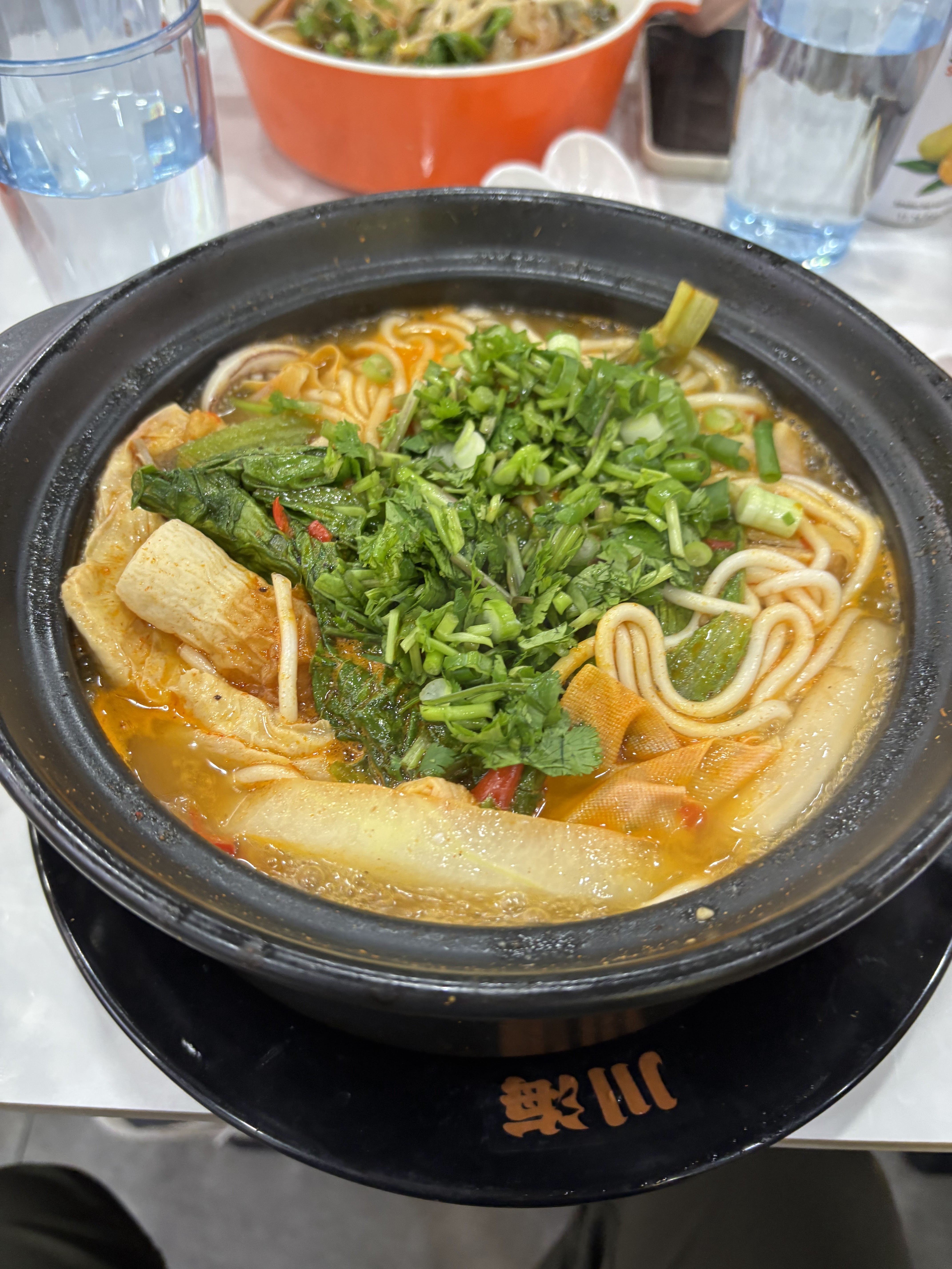 這張圖片中有一碗熱騰騰的湯麵,內有青菜、香菜、豆皮以及辣湯底,看起來非常豐富。桌上還有一杯水與另一份餐點,圖中沒有明顯浮水印。