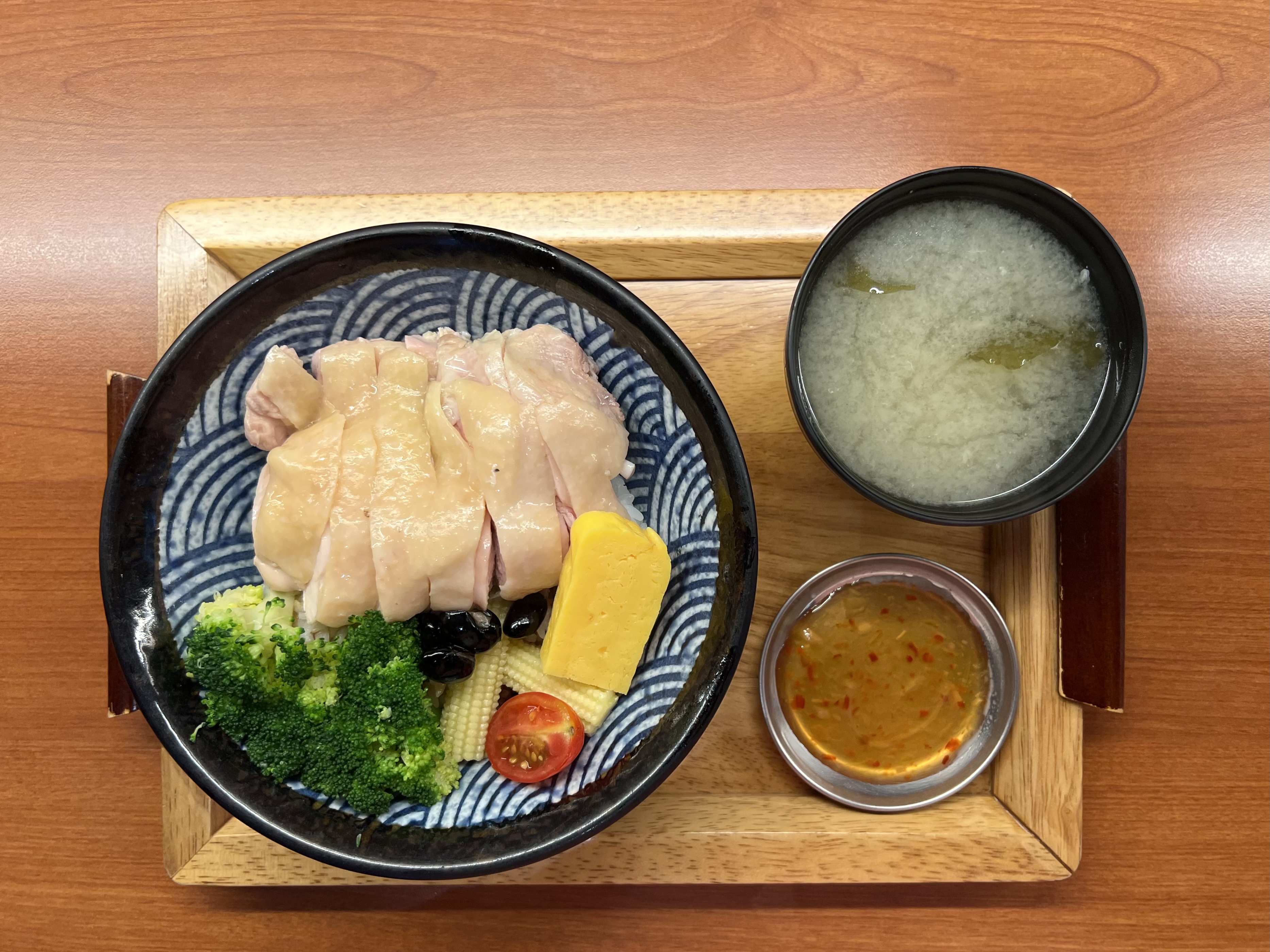 這張圖片是一份雞肉飯便當，主要有白切雞肉、燙青花椰菜、玉米筍、番茄、黑豆、玉子燒，以及一碗味噌湯和一小碟沾醬，擺放於木盤上。圖片無浮水印。