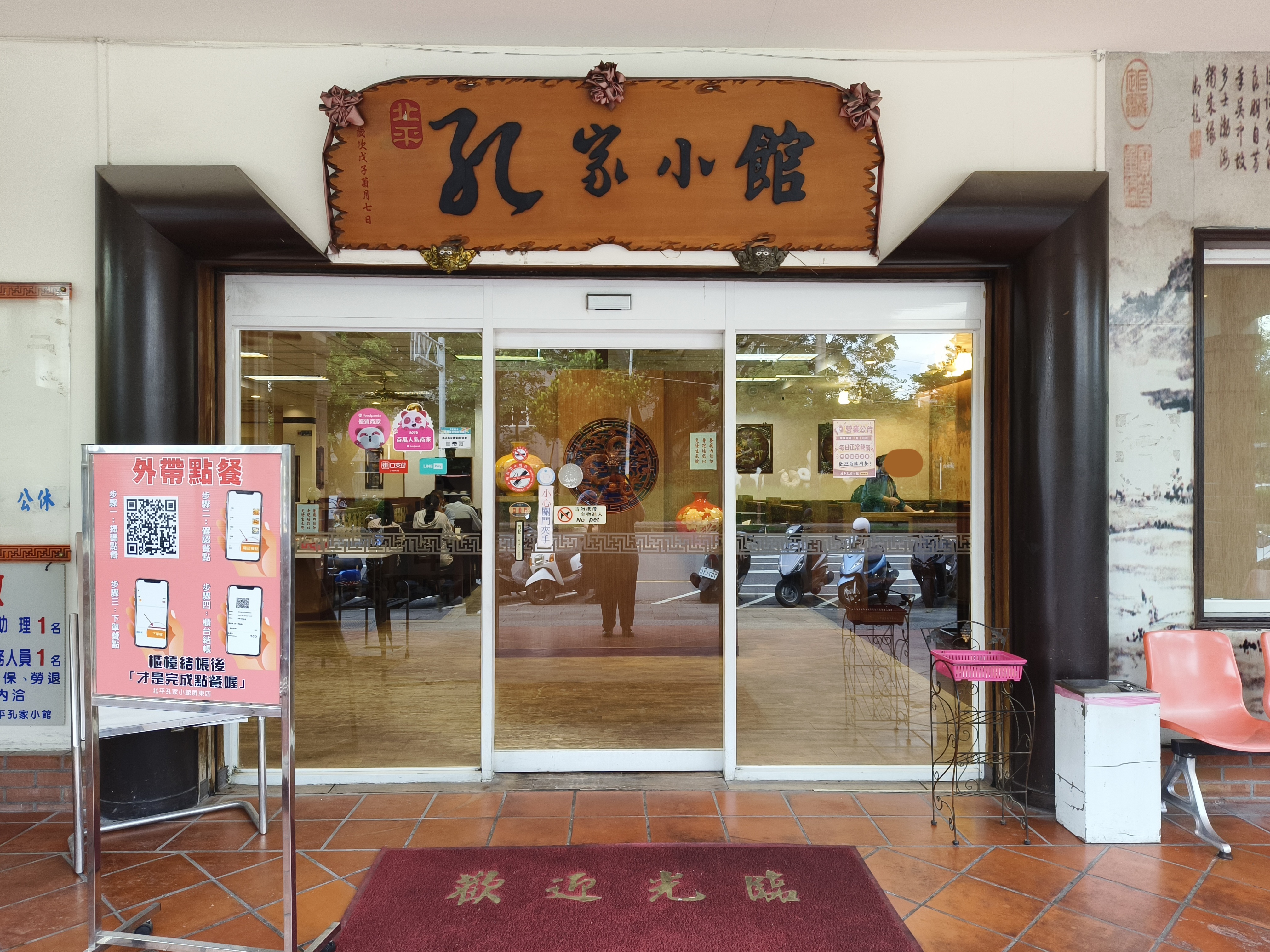這張圖片主要顯示一家名為孔家小館的餐廳入口，有木製招牌、玻璃自動門，以及外帶點餐的告示牌（無浮水印）。門口擺有紅色地墊和座椅，店內可見用餐環境。