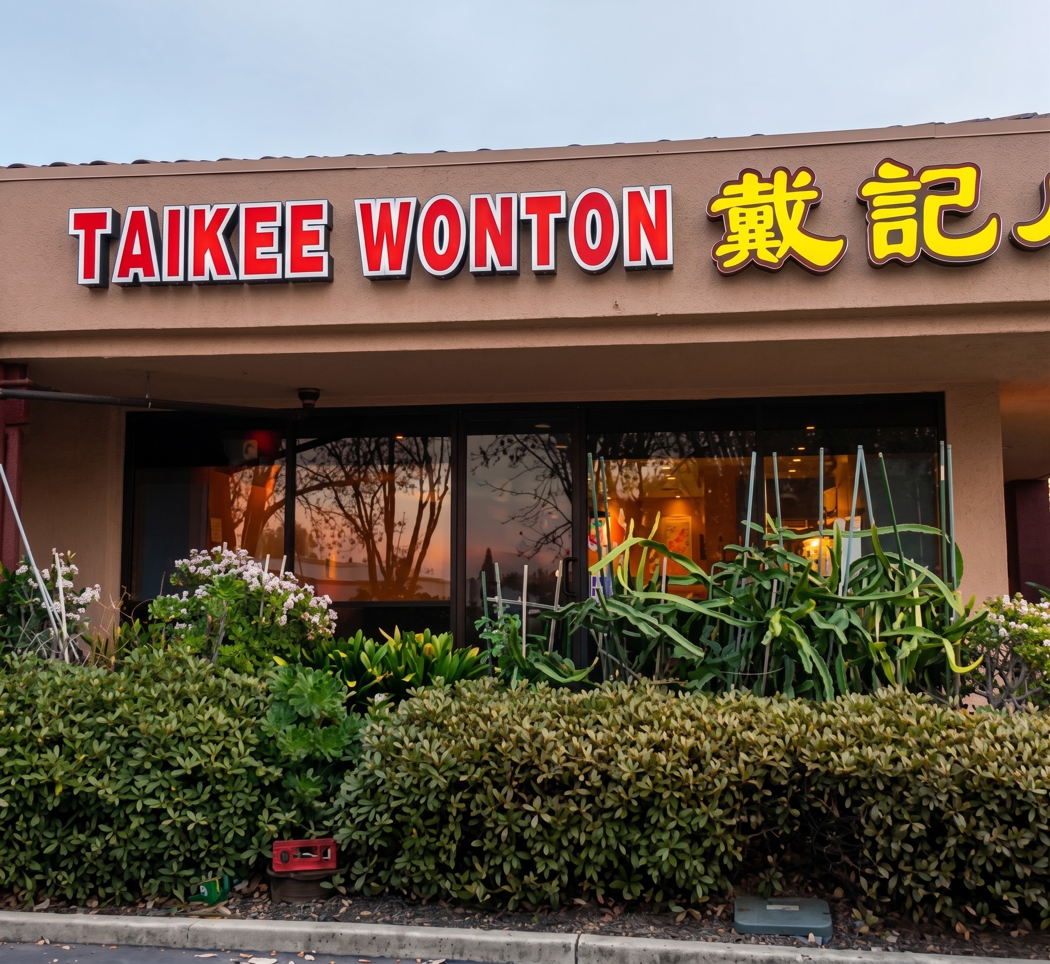 這張圖片主要是一家名為 TAIKEE WONTON 戴記餛飩的餐廳外觀,店門口有綠色植物裝飾,玻璃反射出夕陽與樹木景色。圖片內標誌明顯,無浮水印。