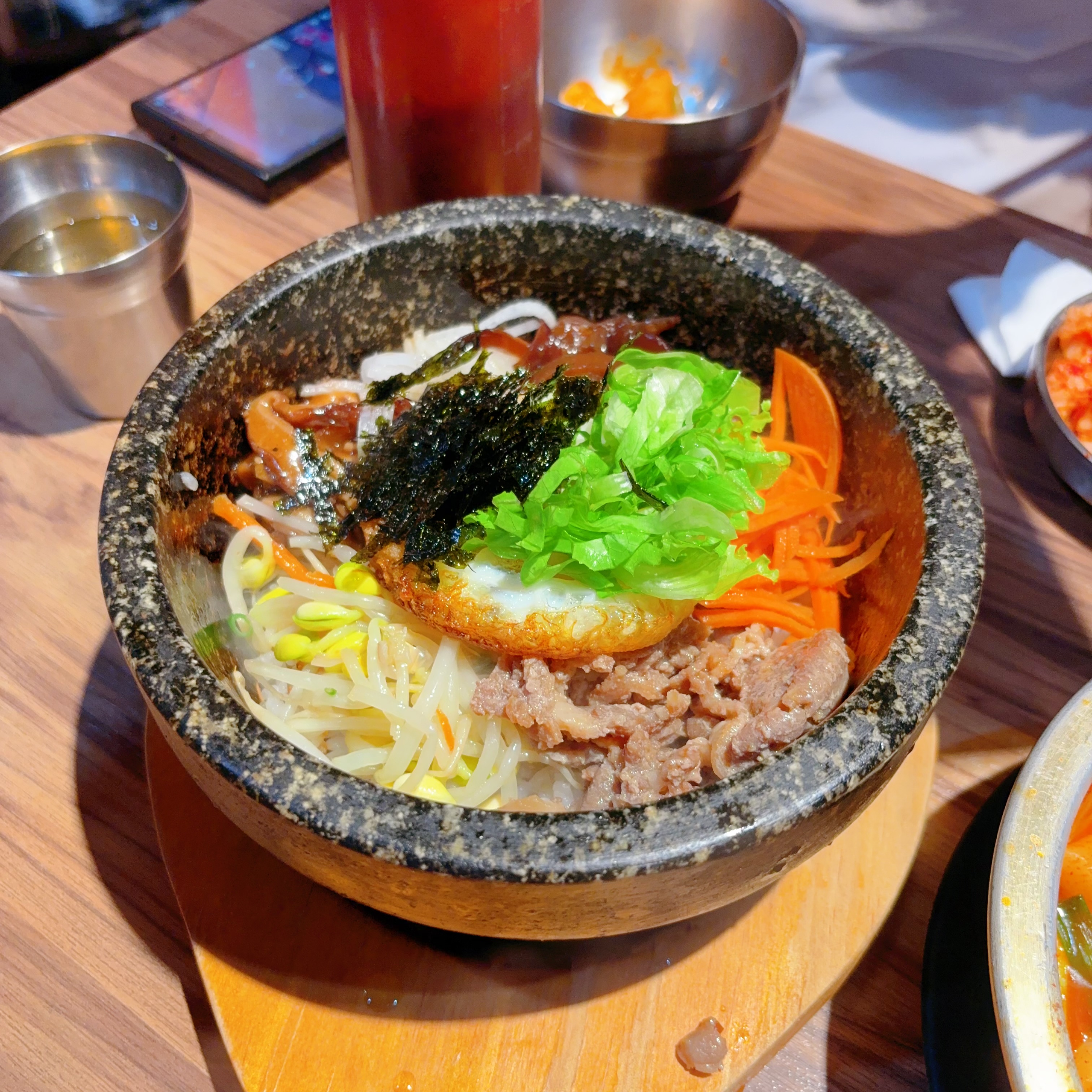 這張圖片主要是一碗韓式石鍋拌飯，碗內有牛肉、荷包蛋、豆芽、紅蘿蔔、海苔、生菜等多樣食材。背景有金屬碗和飲料，但沒有浮水印。