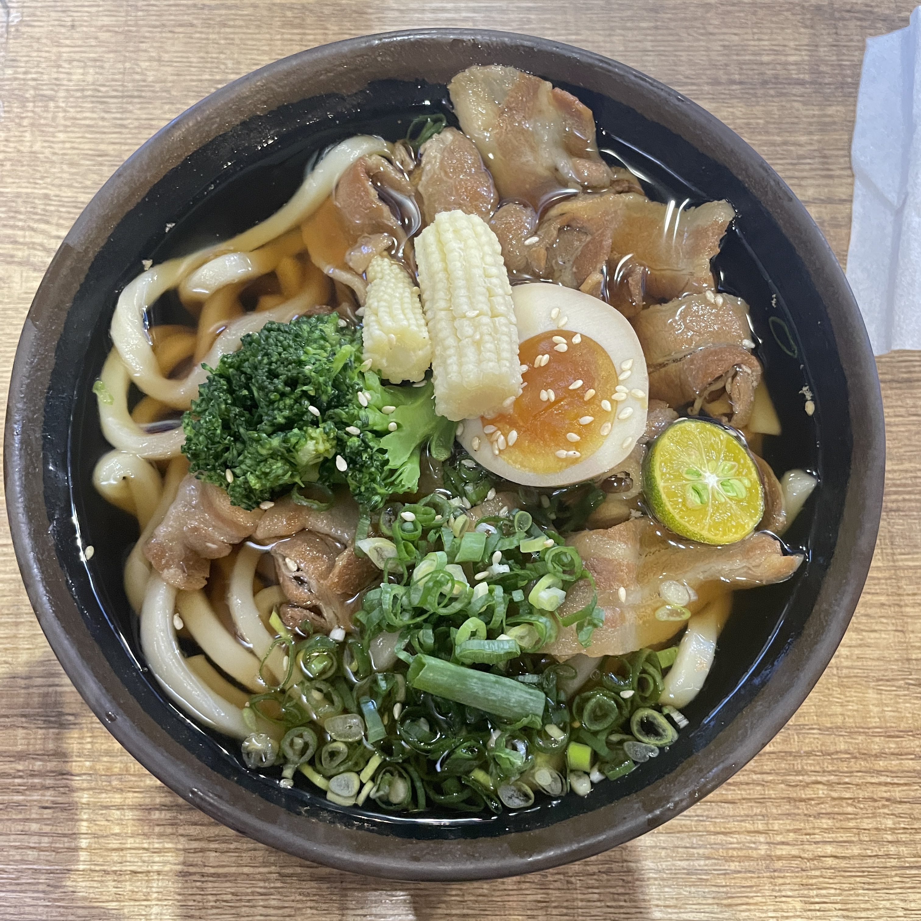 這張圖片是一碗日式烏龍麵，配料有半顆溏心蛋、玉米筍、青蔥、花椰菜、金桔和多片滷豬腳，湯色清澈，擺盤豐富且美觀。圖片無明顯浮水印。