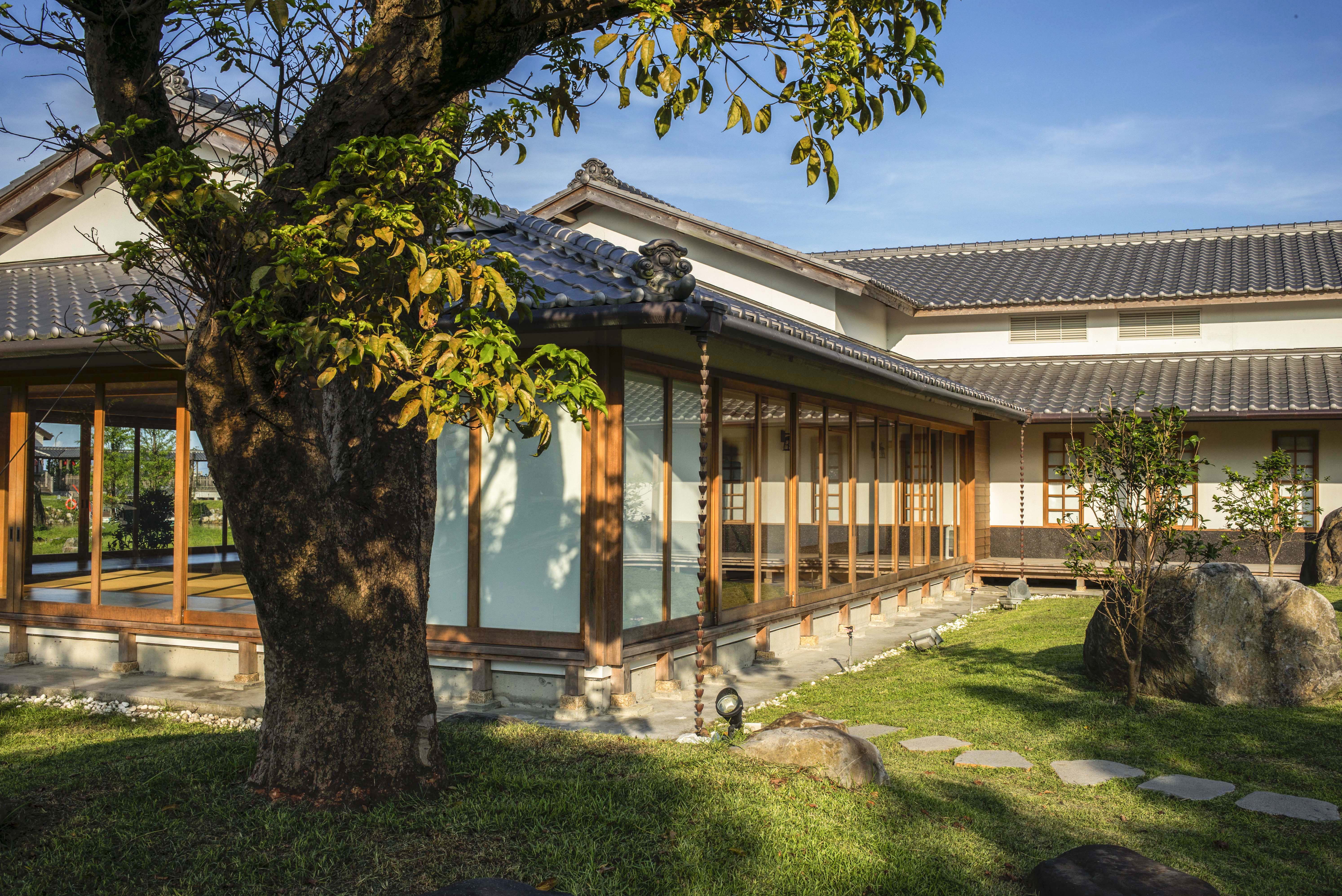 這張圖片展示一幢傳統日式建築,擁有木質結構與拉門,周圍有綠意盎然的庭院、大樹和石頭步道,營造出寧靜自然的氛圍。圖片上沒有浮水印。
