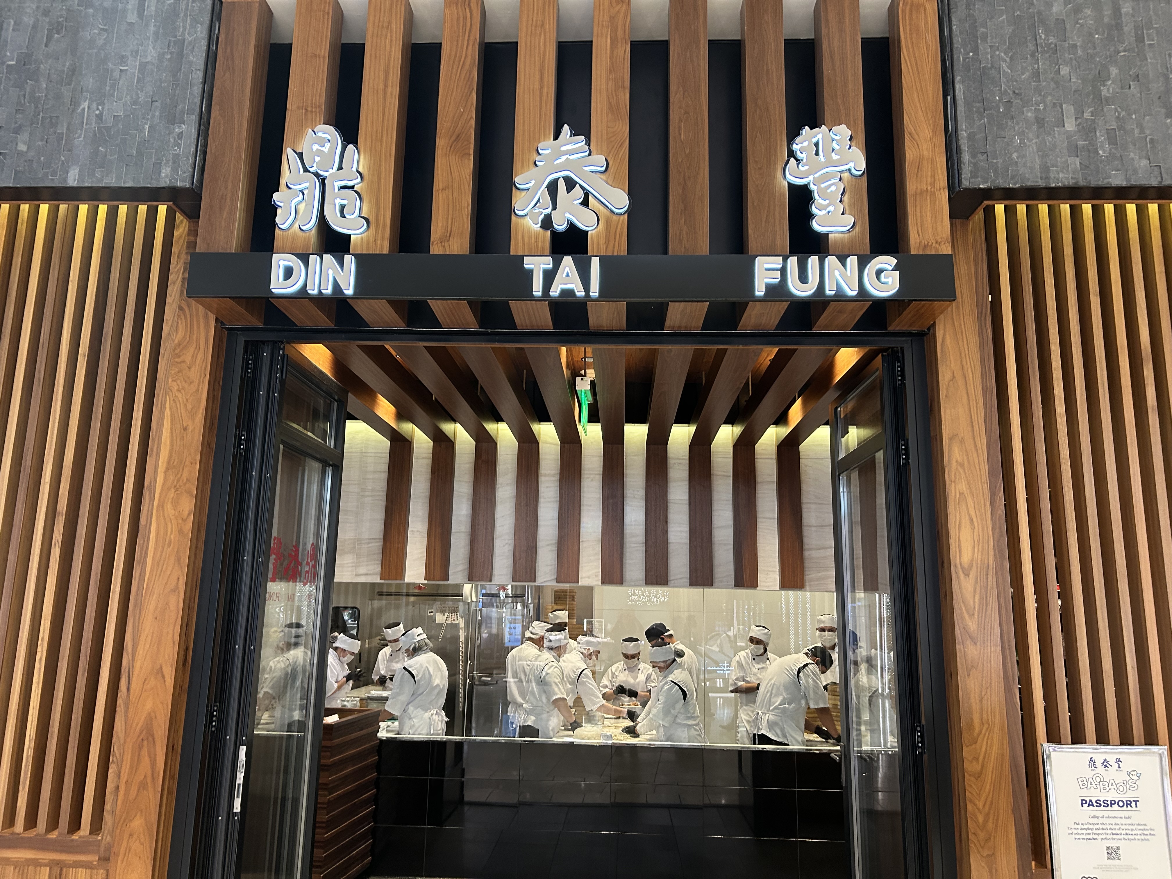 這張圖片主要顯示鼎泰豐餐廳的正門,招牌明顯,內部有多位廚師正在製作點心,現場氣氛專業且整潔。圖片右下角有一張角落立牌,無明顯浮水印。