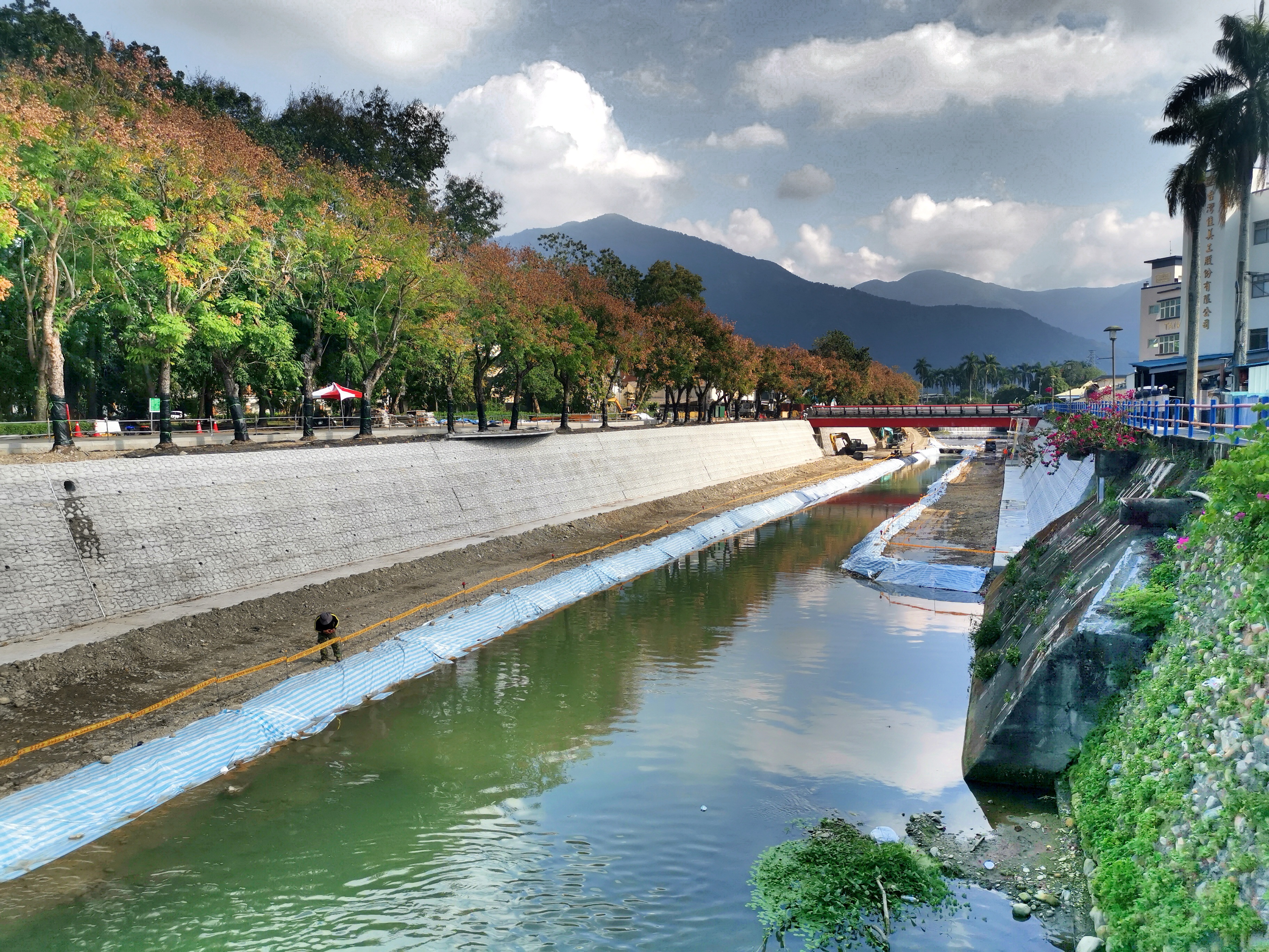 這張圖片主要是一條河川及其兩側的河堤,左側有整齊的綠樹,右側則有建築與花卉。遠方可以看到山脈和藍天,整體環境自然且舒適。圖片上沒有明顯浮水印。