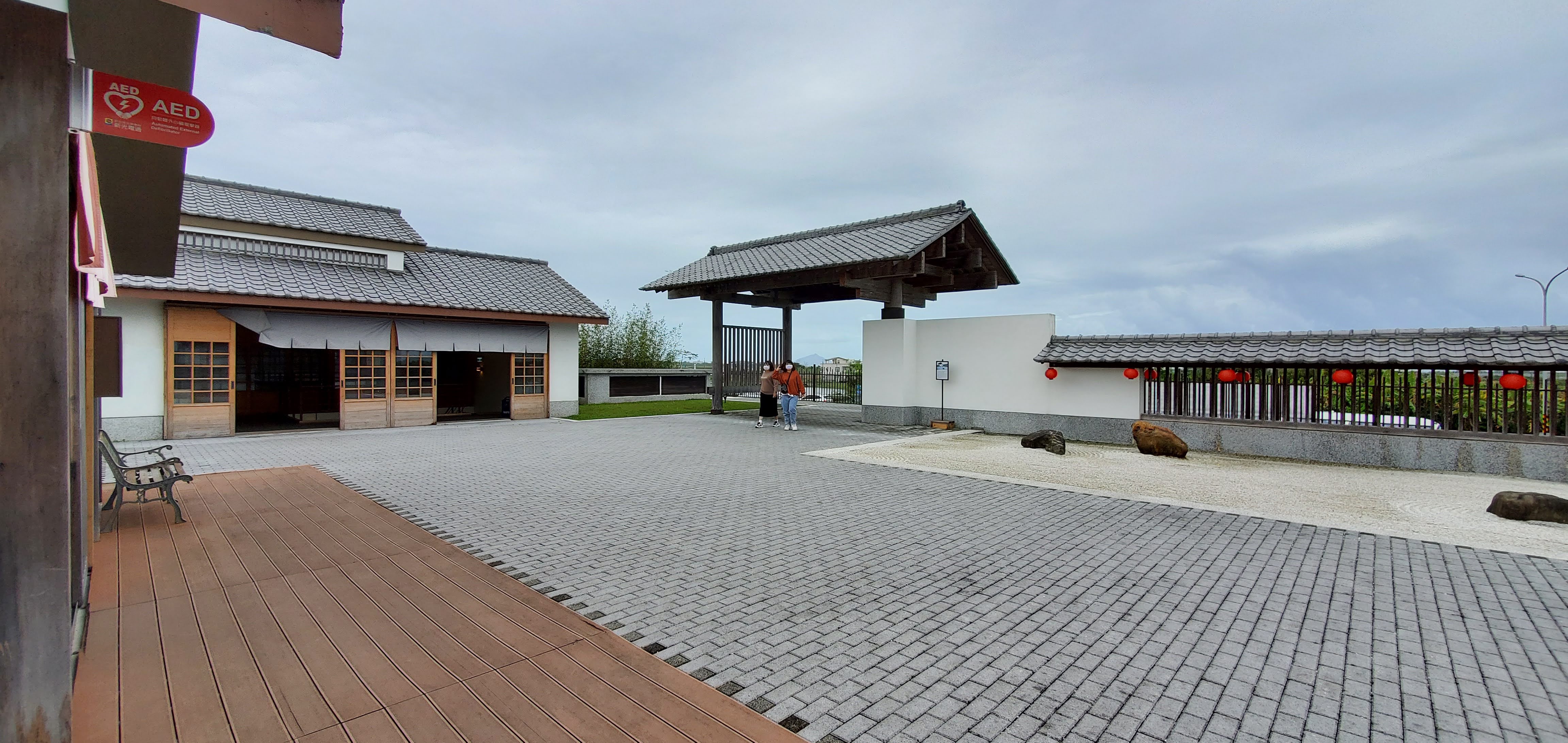 這張圖片主要是一處具有日式建築風格的庭院空間,包含木造建物、拱門和石子地,右側有紅色燈籠裝飾,並有數塊石頭點綴,兩人站於拱門下。左上有AED標誌,無明顯浮水印。