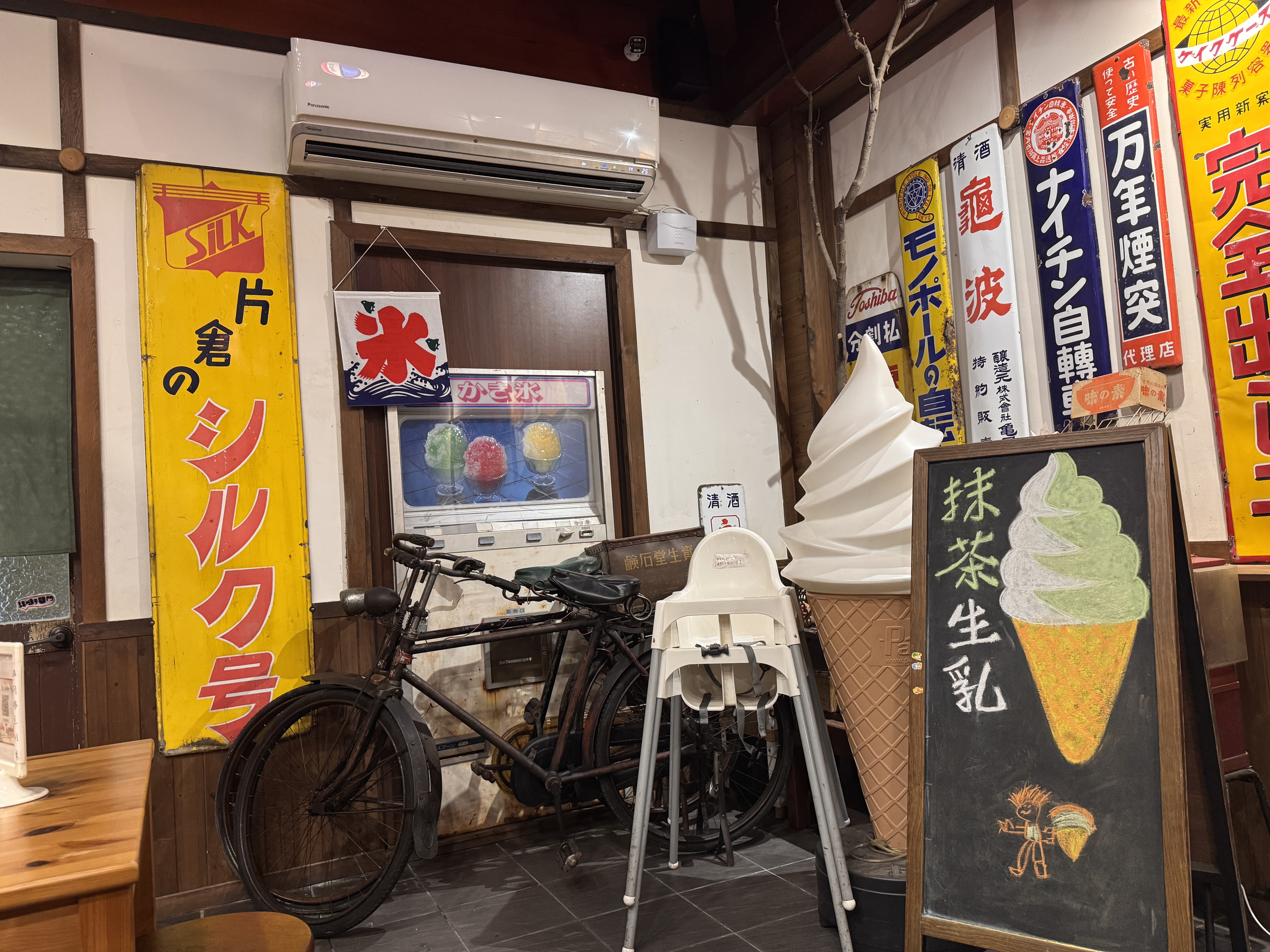 這張圖片展現一家具有懷舊風格的日式店面，牆上有許多復古廣告招牌及刨冰機，還有一台復古腳踏車、冰淇淋模型和寫有抹茶生乳冰淇淋的黑板。圖片未見浮水印。