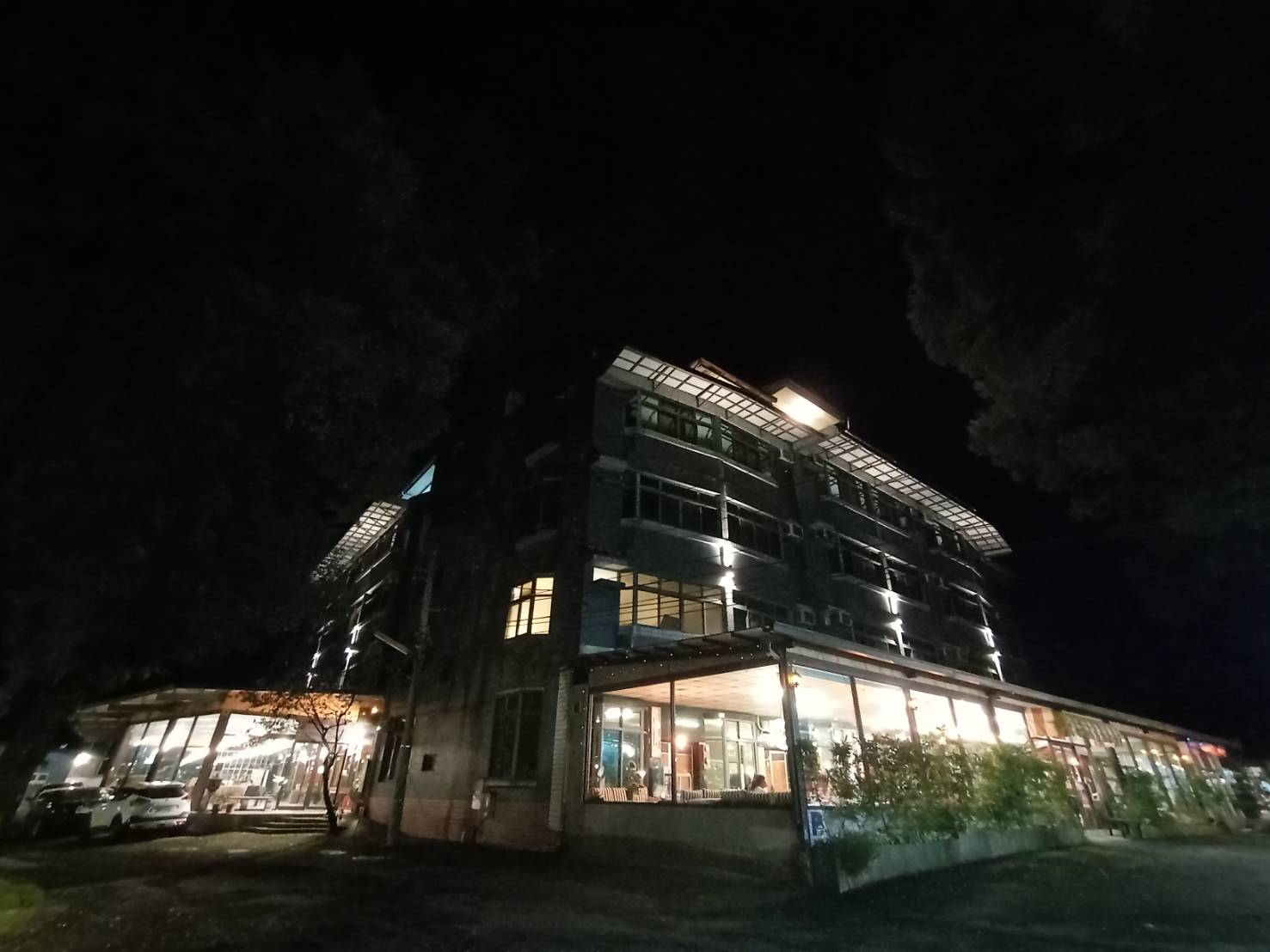 這張圖片主要呈現夜晚時分的一棟大型建築,建築物外觀明亮,有多層樓且玻璃窗戶透出燈光。左側有汽車停放,前方有一些植栽。圖片未見浮水印。