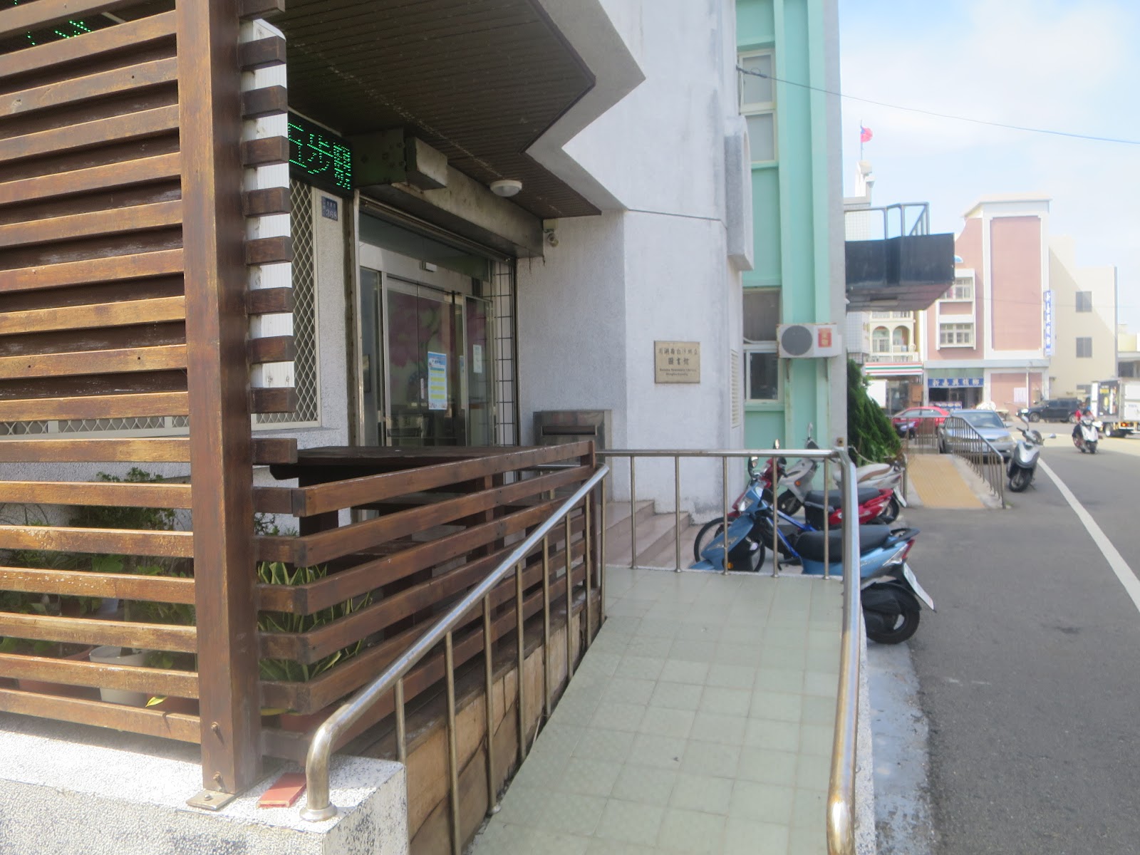圖書館木質格柵入口，門口停著幾輛機車，是在地人借書的日常風景