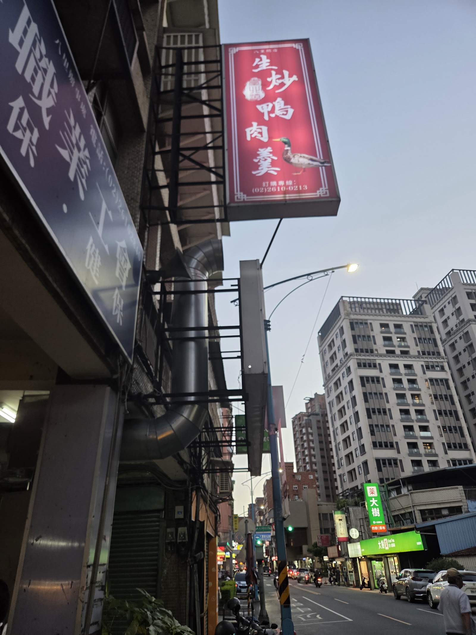臺北港生炒鴨肉羹店家招牌紅底立牌,街景與遠方大樓