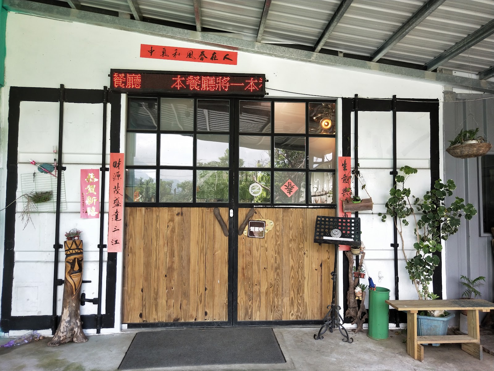 巴奇克竹坊白色鐵皮店面，木拉門搭配黑框玻璃，門口立有木雕圖騰人像，懸掛紅色春聯與LED跑馬燈招牌