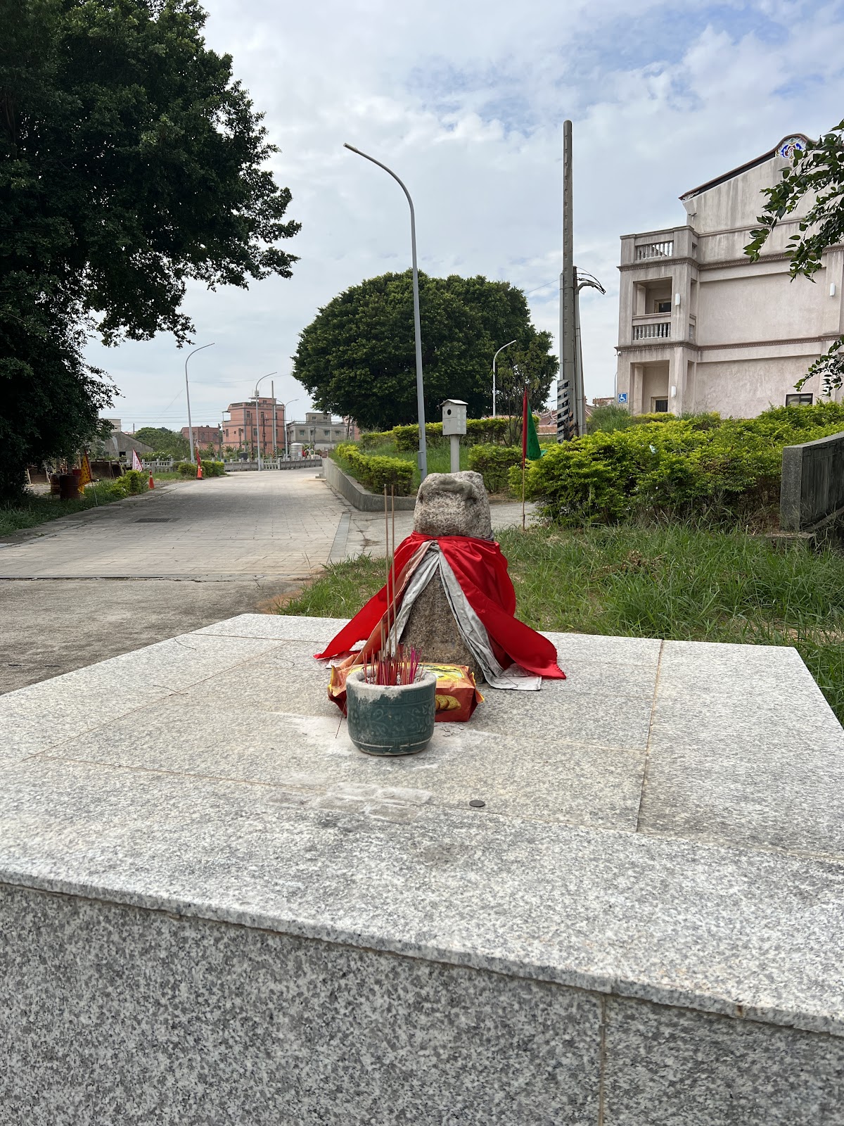 青嶼風獅爺披著紅布端坐村口平台,背景可見僑鄉洋樓與筆直村道