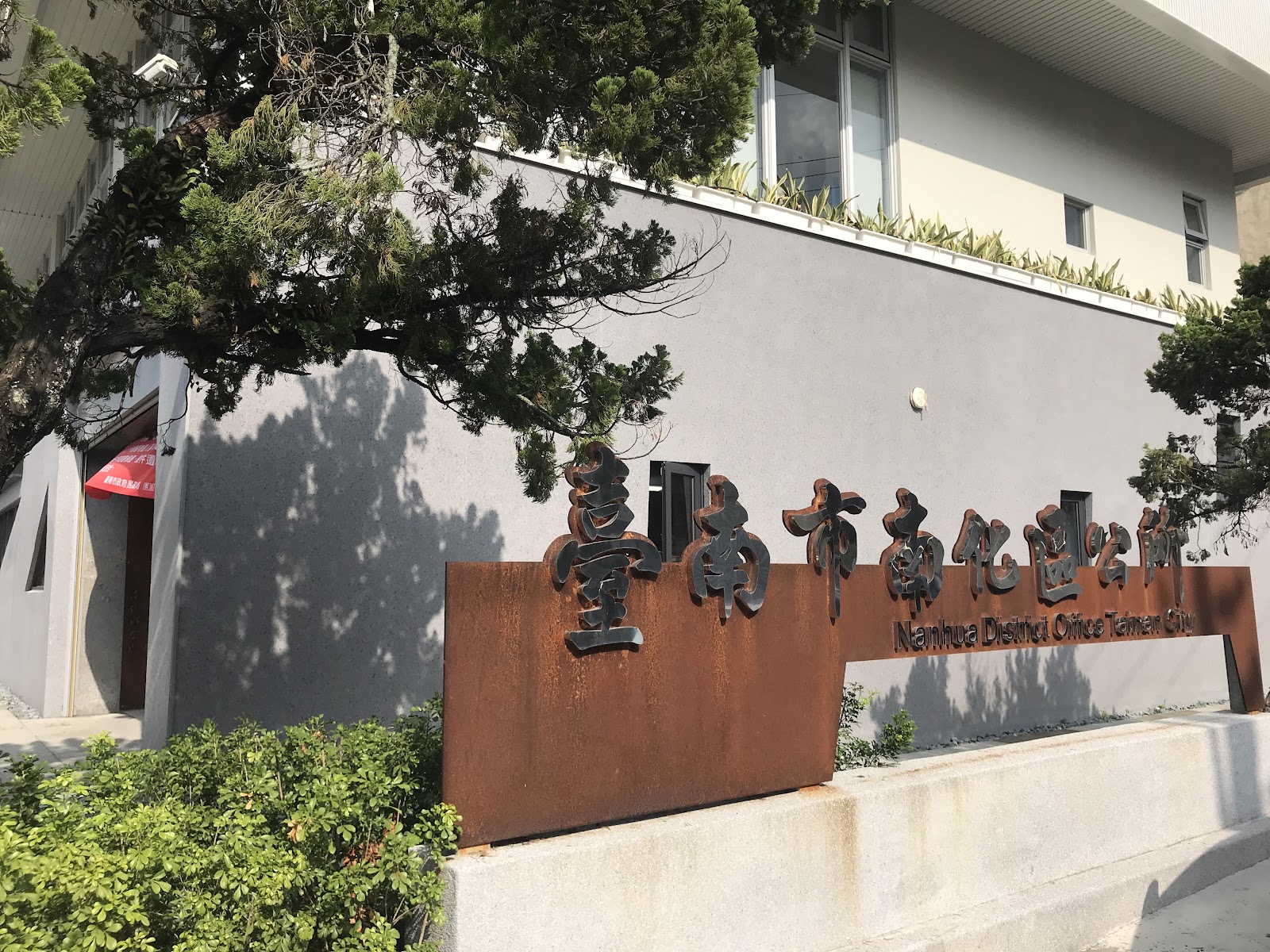 生鏽鋼板材質「臺南市南化區公所」招牌近拍，綠樹灰牆為背景