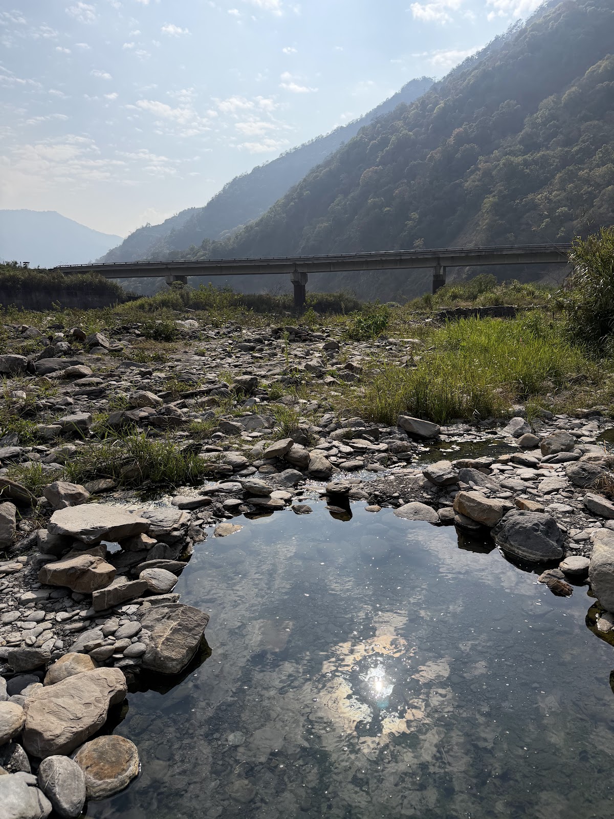 天然溫泉池水面倒映天空，周圍卵石河床，遠方橋樑