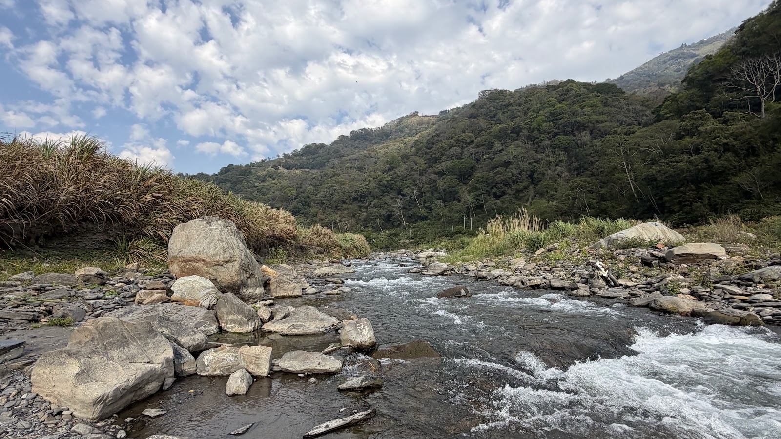 野溪河床遠景，溪水湍急岩石遍布，背景山巒藍天白雲