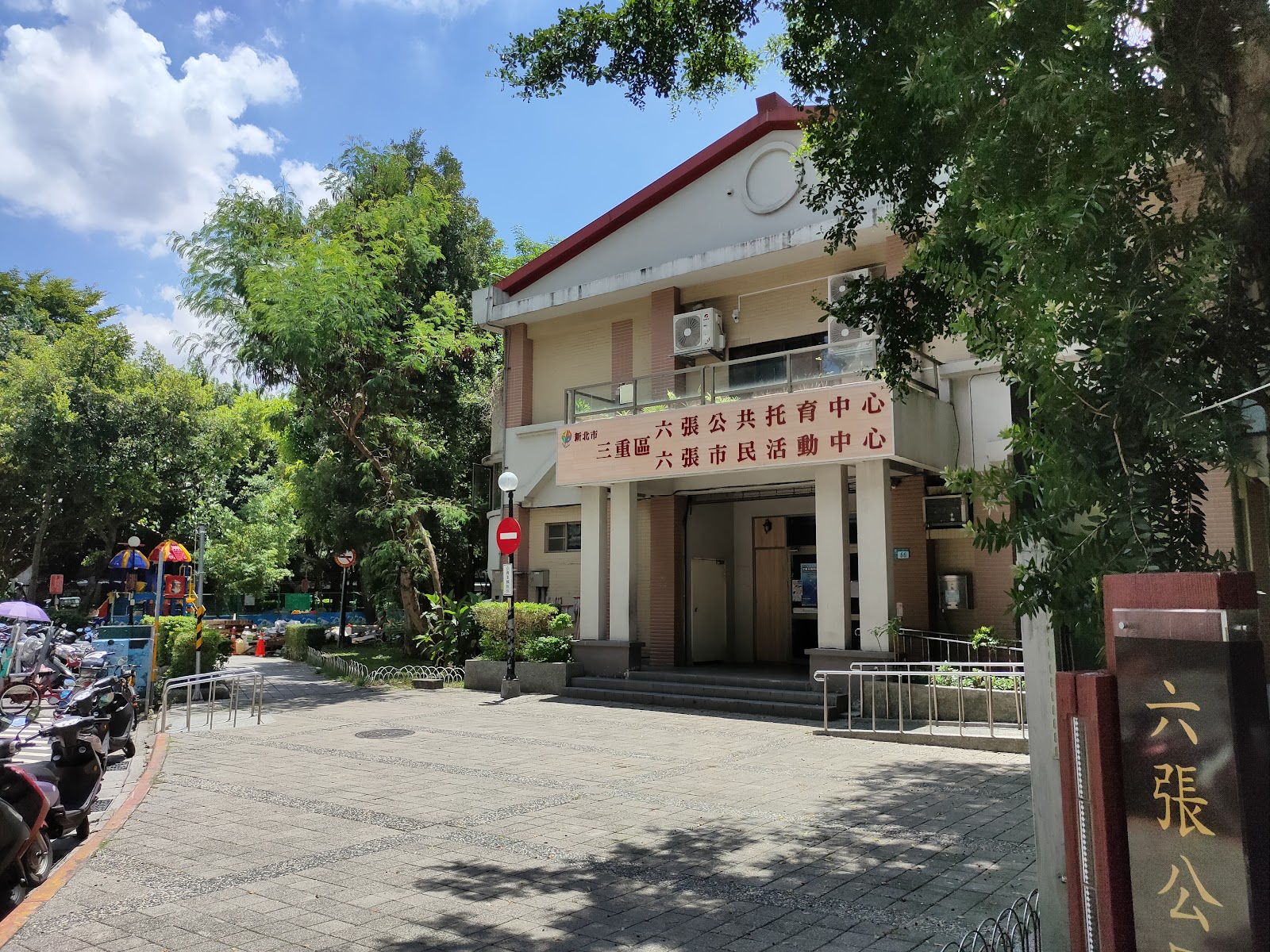 六張公園白天入口石刻地標,後方三重區六張市民活動中心建築物,門口停放多輛機車