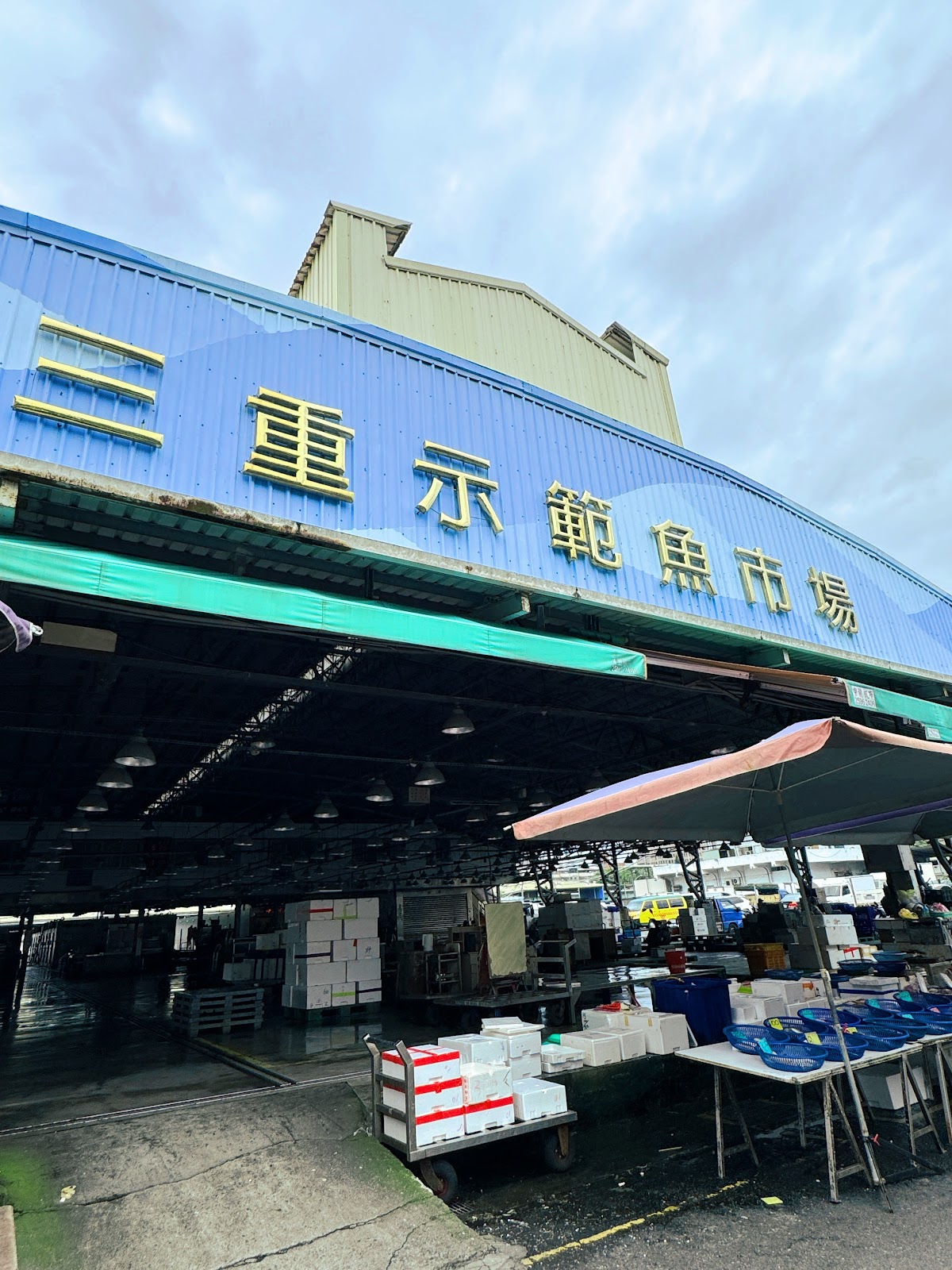 三重力行魚市場藍色鐵皮外觀,黃色大字招牌,下方堆放保麗龍箱與推車