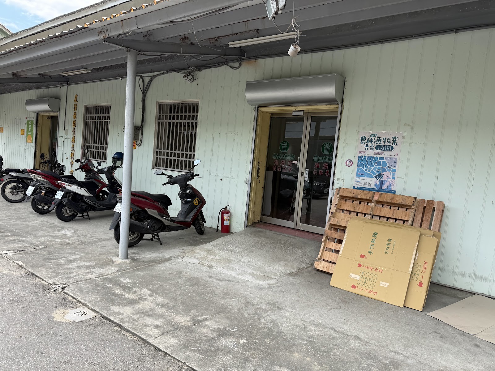 供銷部倉庫式鐵皮建築外觀,騎樓下停放機車與資材紙箱、棧板