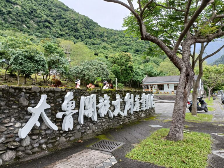 太魯閣族文創園區入口意象牆，石砌矮牆上立體白字招牌搭配傳統服飾人偶雕像，背倚翠綠山巒