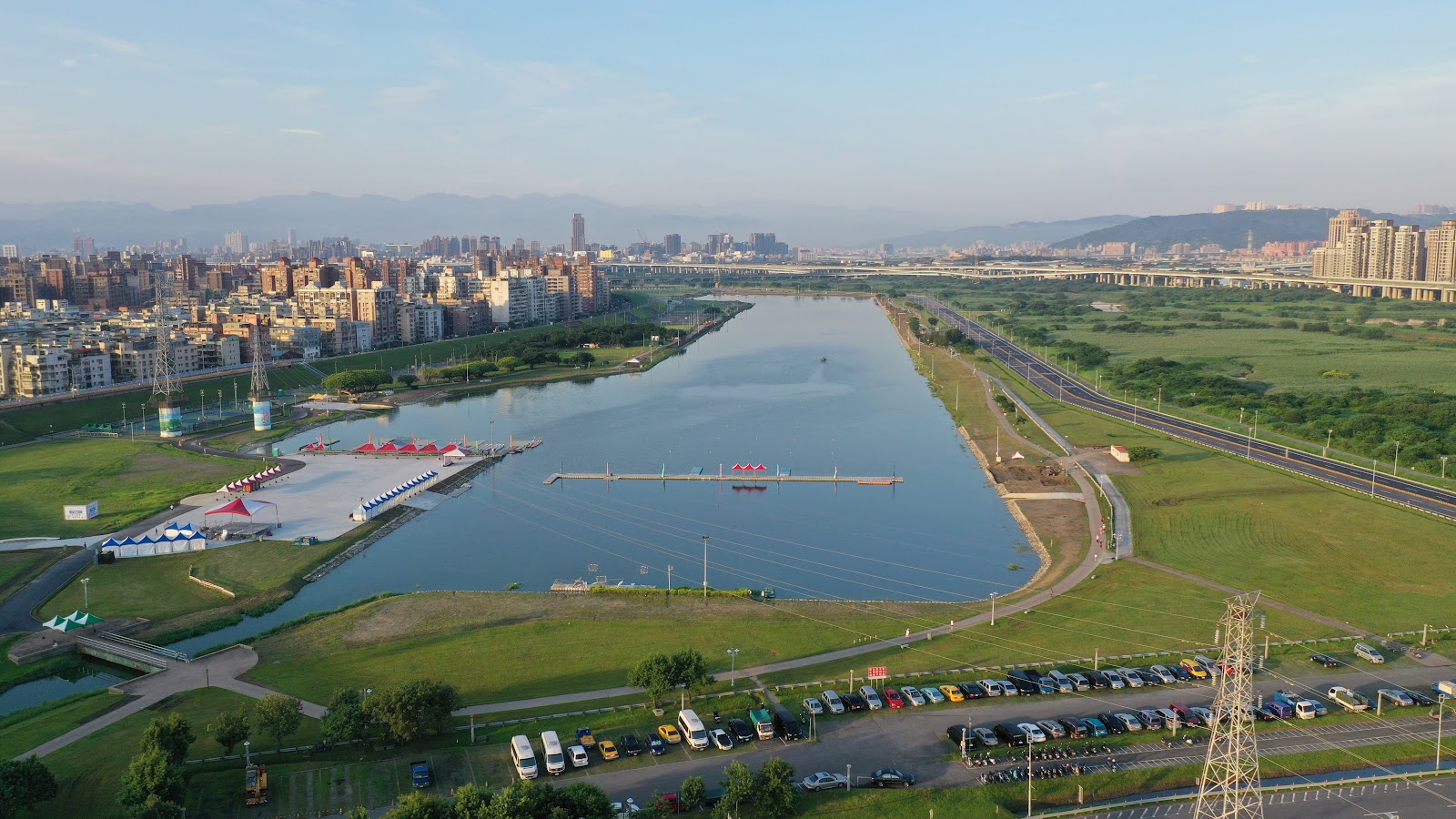 疏洪生態公園空拍水域全景,划船設施紅頂棚、停車場,遠方城市天際線與山巒