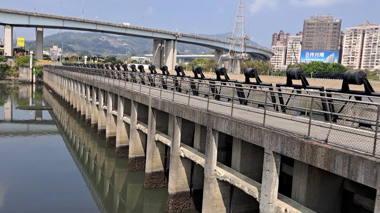 疏洪生態公園水門堤防閘道設施近景,倒影清晰,背景高架橋與城市建築