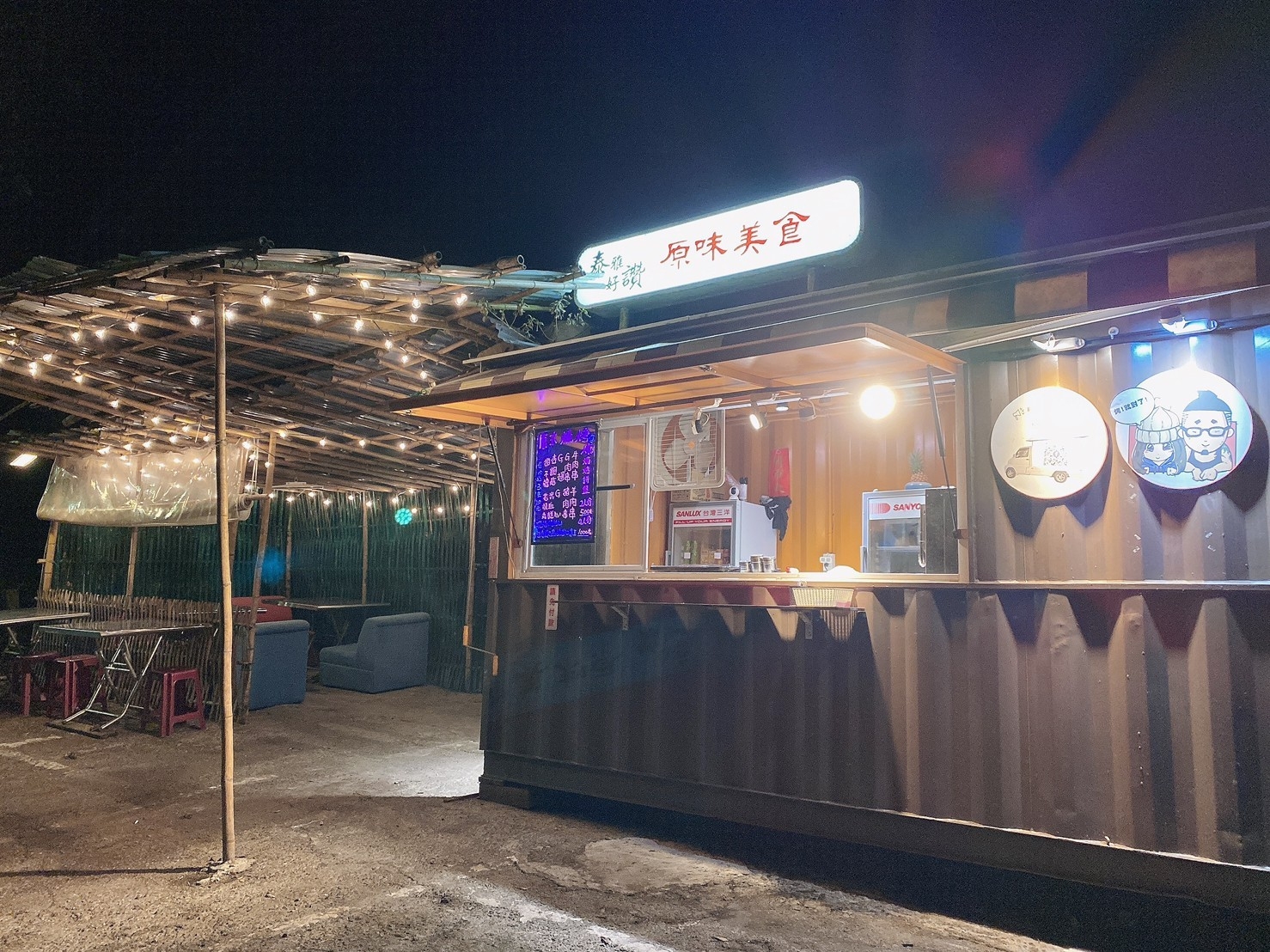 夜晚店門口鐵皮貨櫃攤位，暖黃燈串圍繞竹編棚架